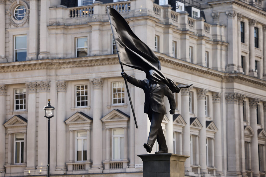 A statue depicting a person stepping off the plinth while carrying a flag covering their face, by British street artist Banksy, stands in London