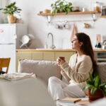 A woman sits quietly on a couch, engaging in deep thought while holding a warm drink.