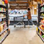 a woman with a cart walks between rows of shelves in a grocery store